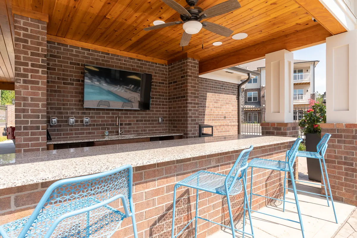 The photo displays a lively outdoor granite bar counter surrounded by blue metal chairs, a mounted flat-screen TV, and brick walls. The area feels energetic and ideal for socializing, watching sports, or hosting gatherings.
