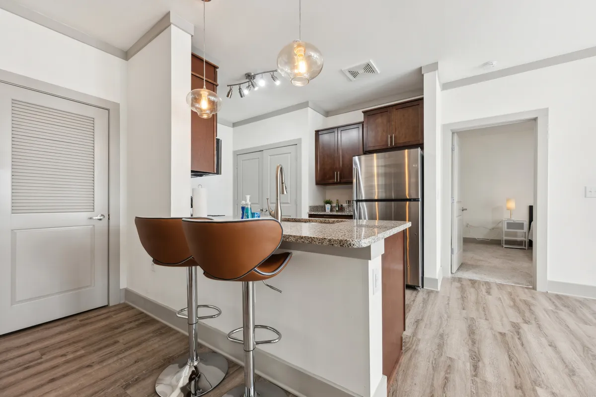 A contemporary kitchen scene featuring a spacious kitchen counter with sleek bar stools, modern cabinetry, and visible appliances. The layout highlights both the cooking area and a space for guests or family to gather, emphasizing a seamless flow between kitchen and casual dining/socializing.