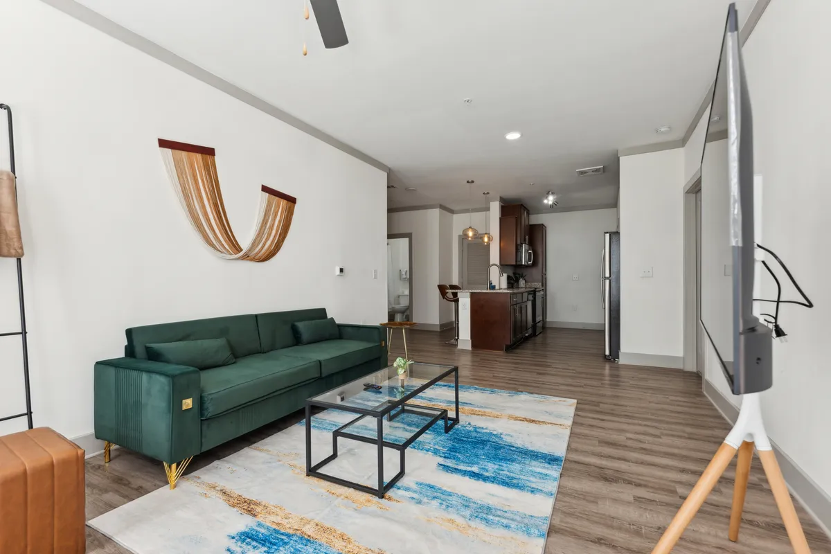 A contemporary living room with a striking green sofa, a glass coffee table, decorative wall art, and a TV stand. The setting exudes comfort and style, with ample natural light and designer touches, emphasizing a space ideal for relaxation or social gatherings.
