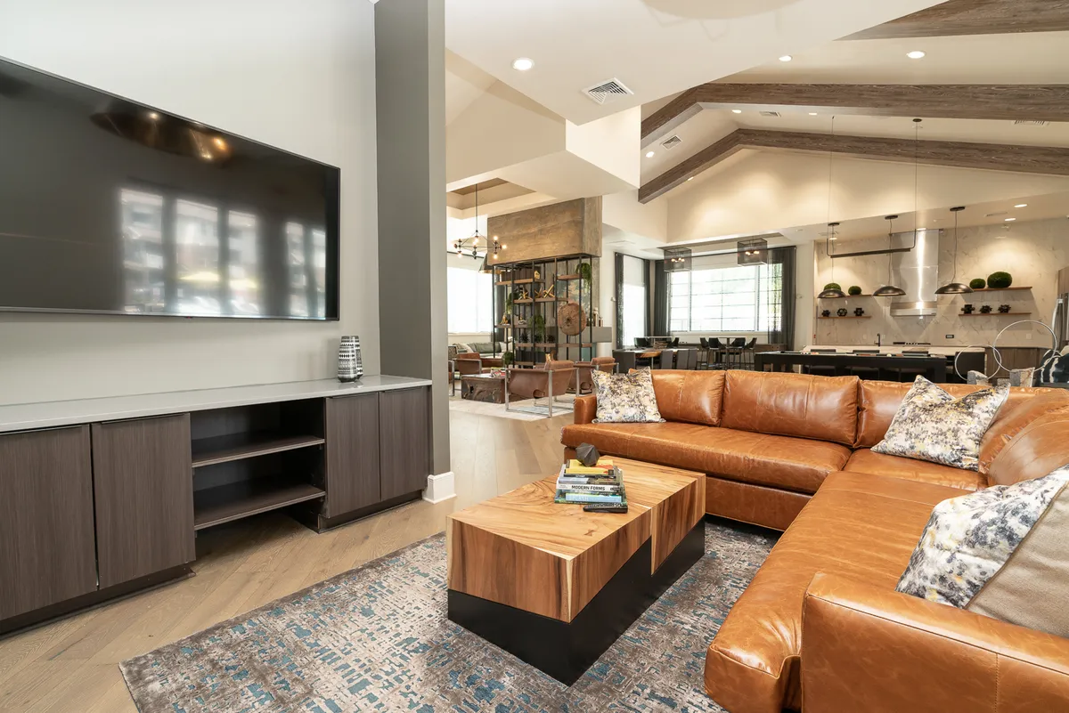 A spacious open-plan living area featuring a large sectional sofa, a stylish coffee table, a television, and a dining area visible in the background. The space is filled with natural light from large windows and is designed with contemporary finishes and thoughtful decor, creating a seamless flow between lounging and dining areas.