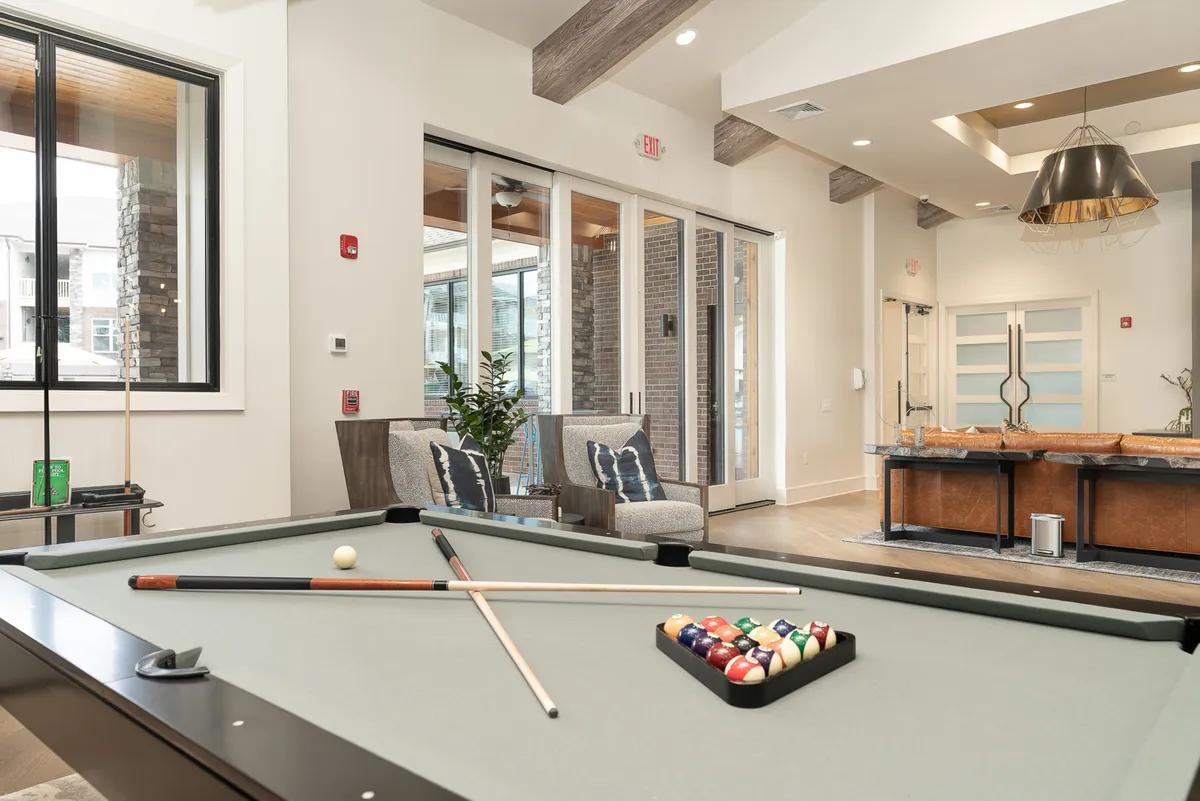 A spacious lounge area featuring a pool table, comfortable lounge chairs, a sleek bar area, and large windows that fill the room with natural light. The setup encourages friendly interaction and leisure while offering a relaxed, upscale vibe.