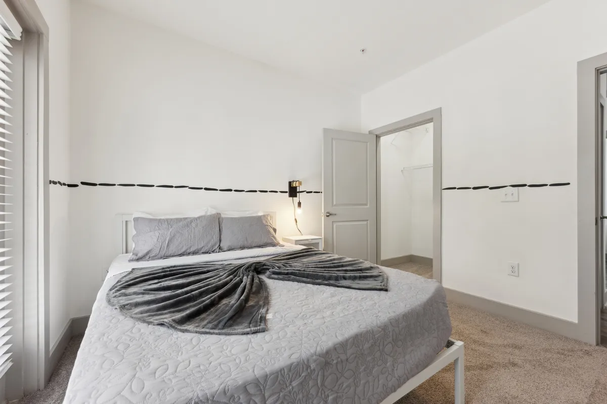 A softly lit bedroom showcasing a bed with stylish gray bedding, a decorative throw blanket, and visible closet space. The room is bathed in natural light, emphasizing tranquility and an inviting atmosphere perfect for rest or quiet reading.